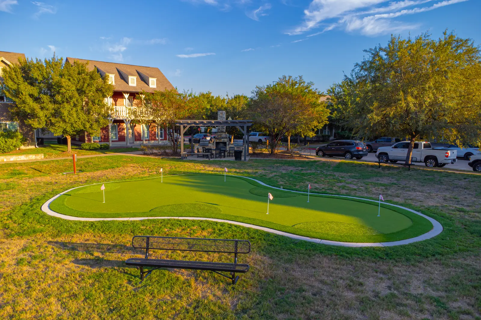 DJI_0818 Putting green with several practice holes, a bench, perimeter fencing, and nearby apartments